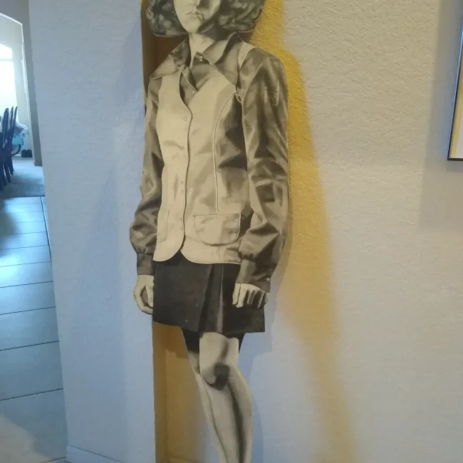 Life-size pencil drawing of the artist when she was 17 on a plywood stand. Completely in gray pencil, the artist is depicted with neck-length hair, big round sunglasses on her head, a long collared shirt and buttoned vest, a skirt and airy, heeled shoes.
